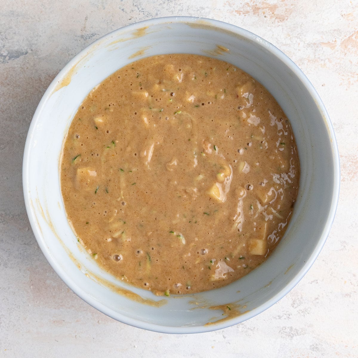 The fully mixed apple zucchini bread batter in a ceramic mixing bowl. The batter is light brown with chunks of apple and flecks of green from the zucchini peel