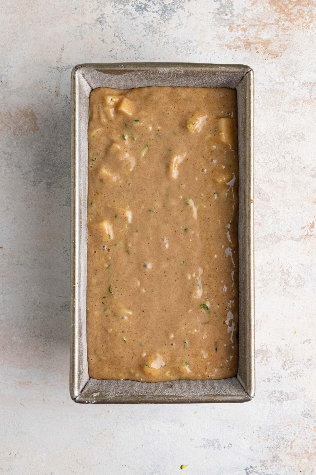 The apple zucchini bread batter in a 8.5x4.5-inch metal loaf pan. The batter is light brown with flecks of green from the zucchini peel.