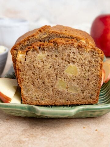A close-up of a sliced loaf of apple zucchini bread on a white plate, showing a moist texture with apple chunks and small bits of zucchini visible throughout.