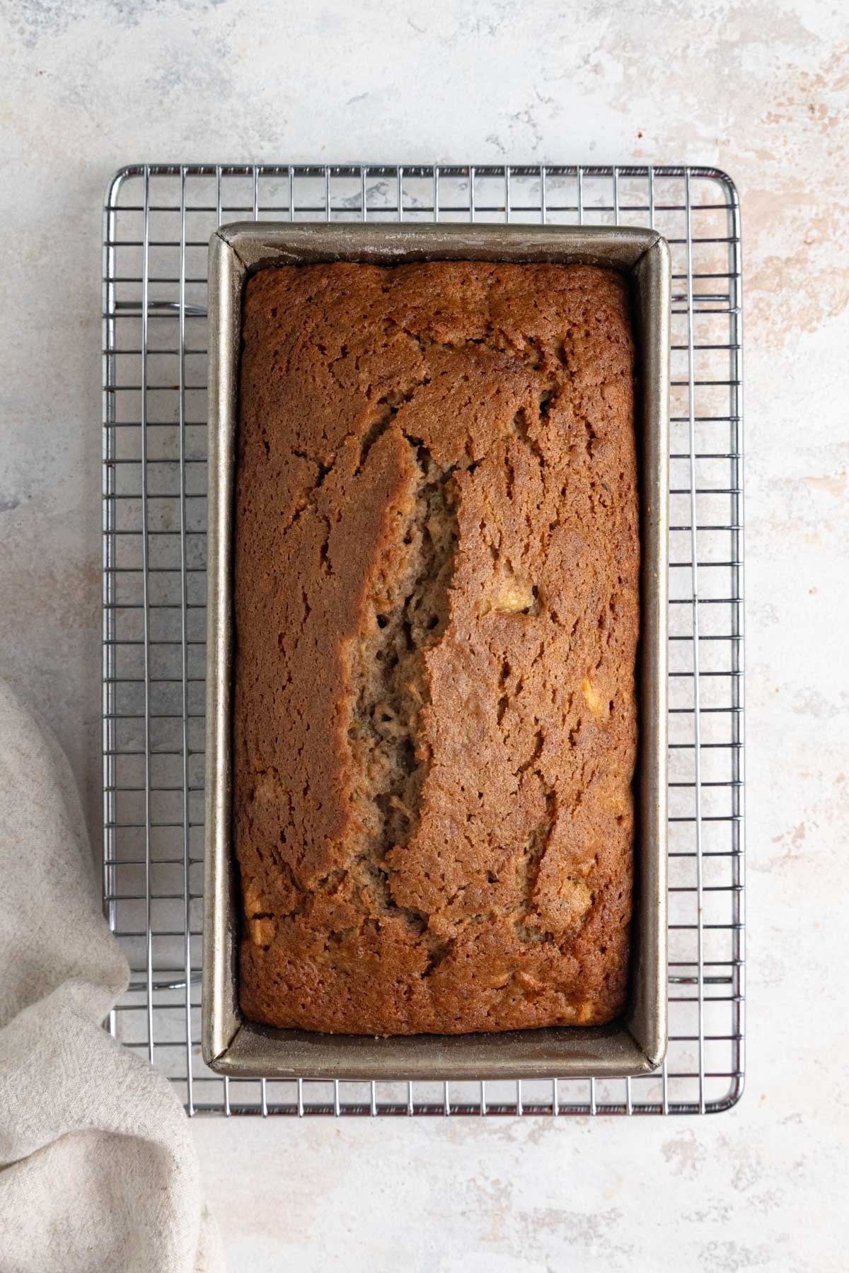 A loaf of apple zucchini bread sits in a rectangular metal loaf pan on a wire cooling rack.
