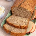 A loaf of freshly baked zucchini bread is partially sliced on a white platter with a wooden knife. A small bowl of butter and a cup of tea are nearby, ready for serving. One slice of the moist apple zucchini bread is buttered on a separate plate.
