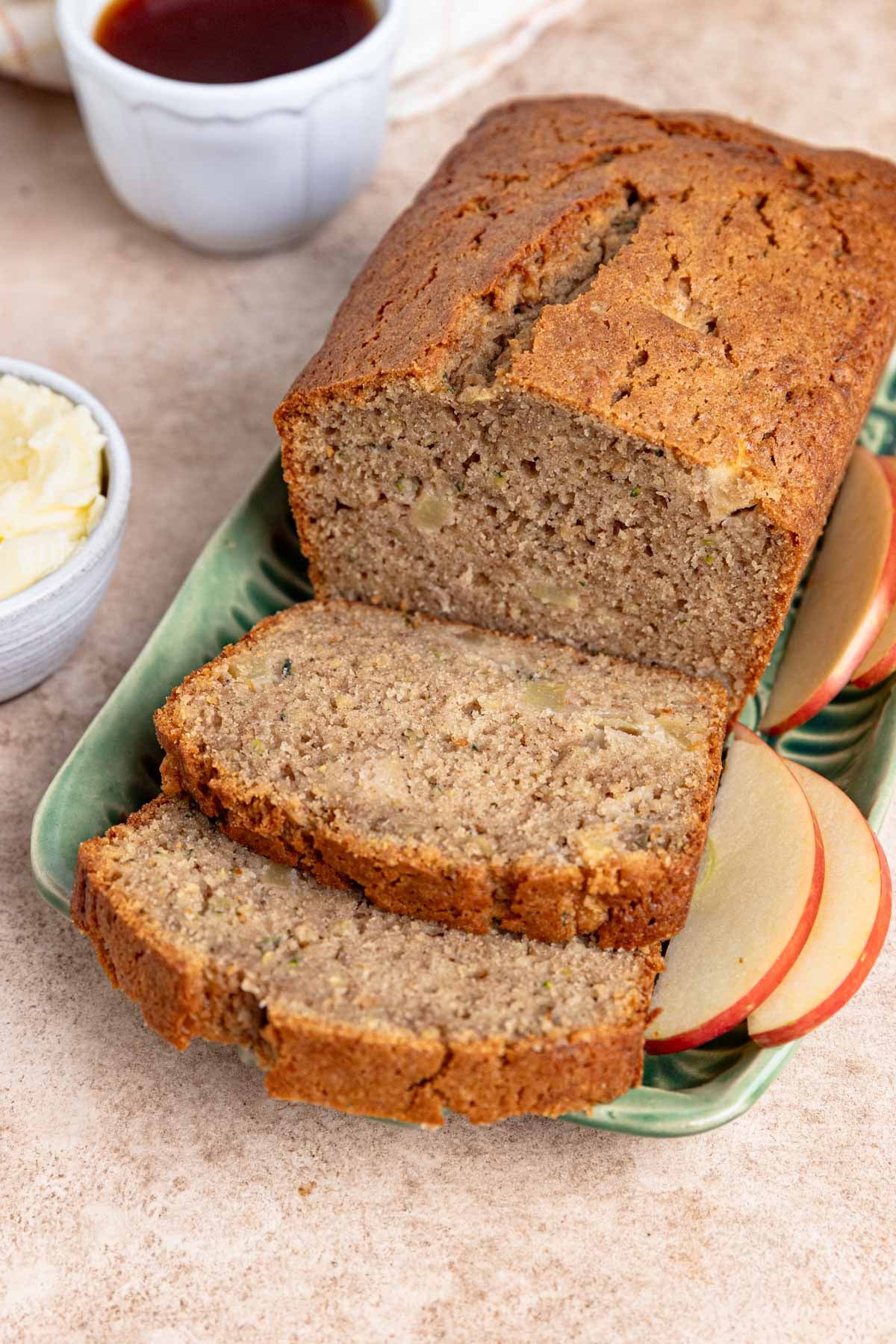 A loaf of freshly baked zucchini bread is partially sliced on a white platter with a wooden knife. A small bowl of butter and a cup of tea are nearby, ready for serving. One slice of the moist apple zucchini bread is buttered on a separate plate.