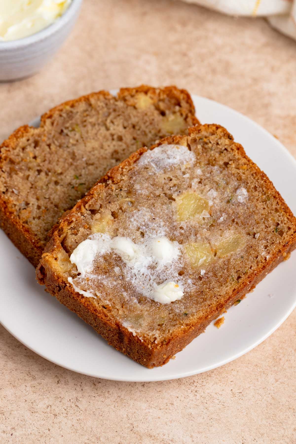 A slice of apple zucchini bread with butter spread on top sits on a white plate. A bowl of butter are visible in the background.