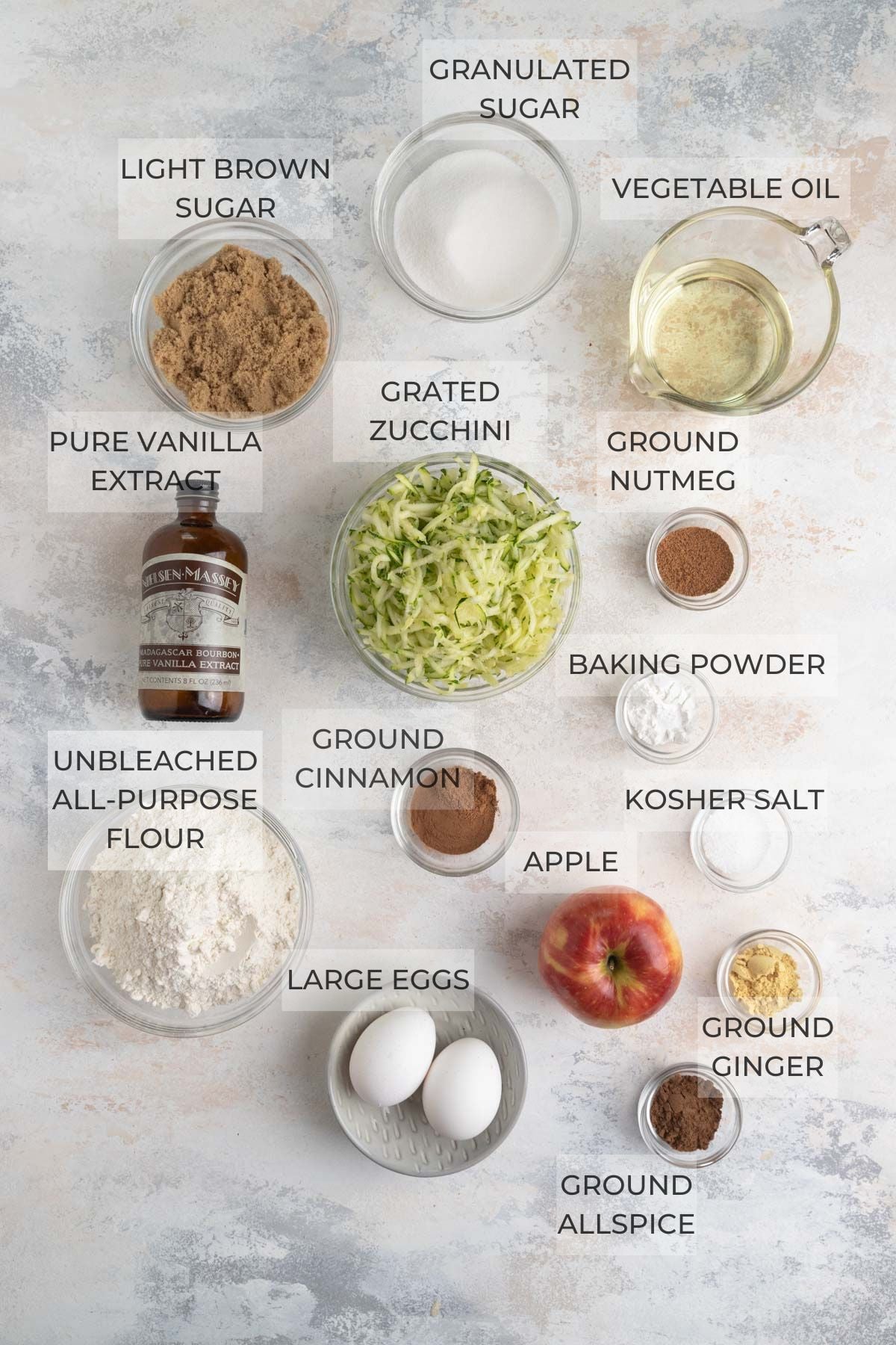 Top-down view of labeled baking ingredients in bowls and containers, including granulated sugar, vegetable oil, unbleached all-purpose flour, pure vanilla extract, grated zucchini, baking powder, ground cinnamon, ground nutmeg, ground cloves, kosher salt, and eggs.