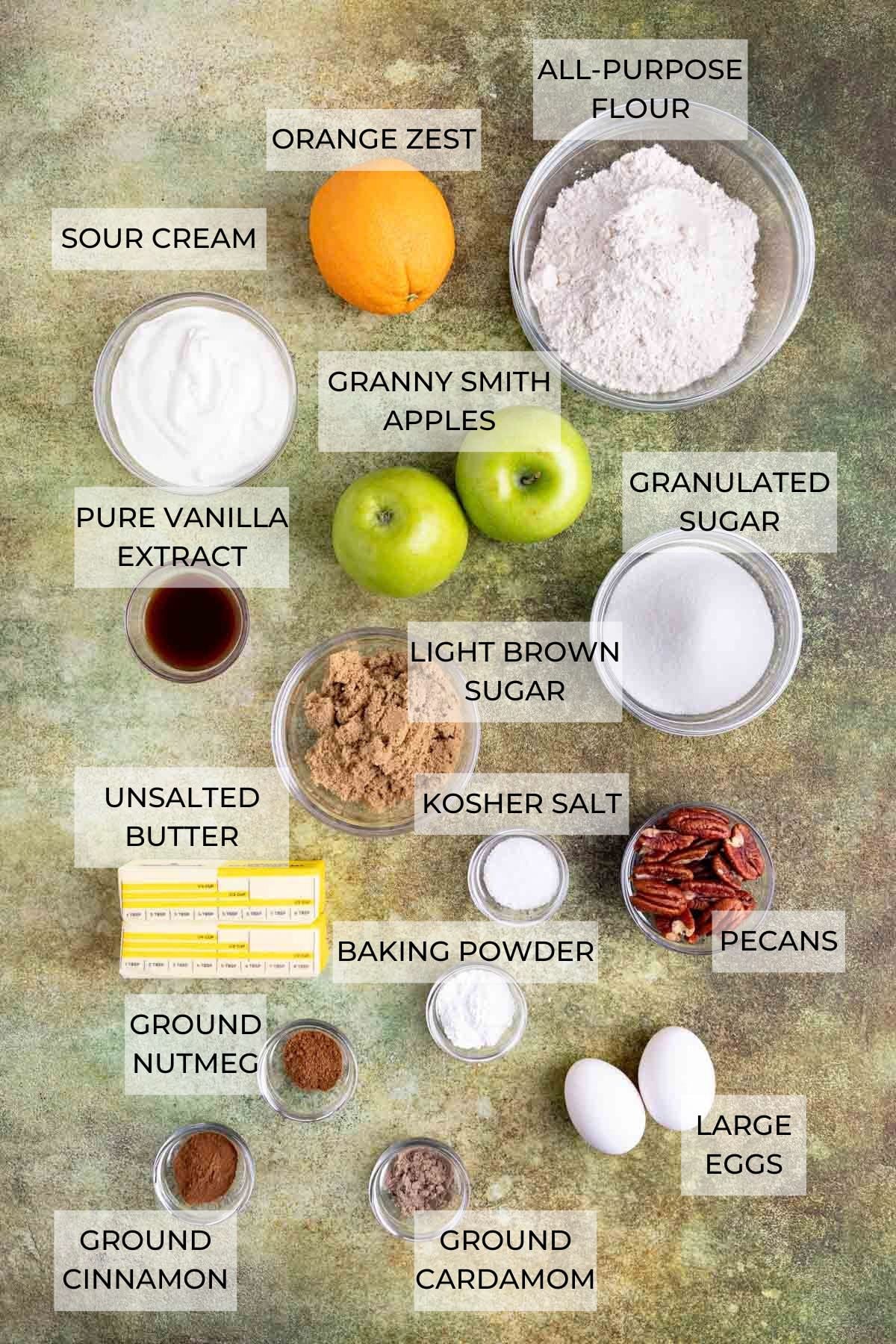 Apple streusel muffin ingredients arranged on a green backdrop. Ingredients include an orange, all-purpose flour, granulated sugar, pecans, eggs, kosher salt, baking powder, cinnamon, nutmeg, cardamom, unsalted butter, brown sugar, pure vanilla extract, Granny Smith apples, and sour cream