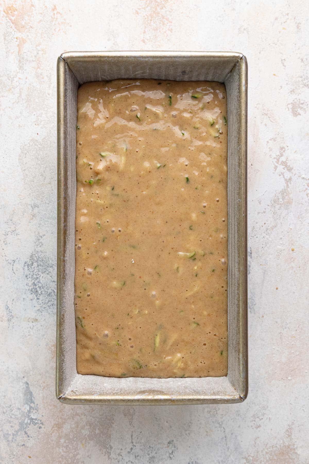The zucchini bread batter in a 8.5x4.5-inch metal loaf pan. The batter is light brown with flecks of green from the zucchini peel.