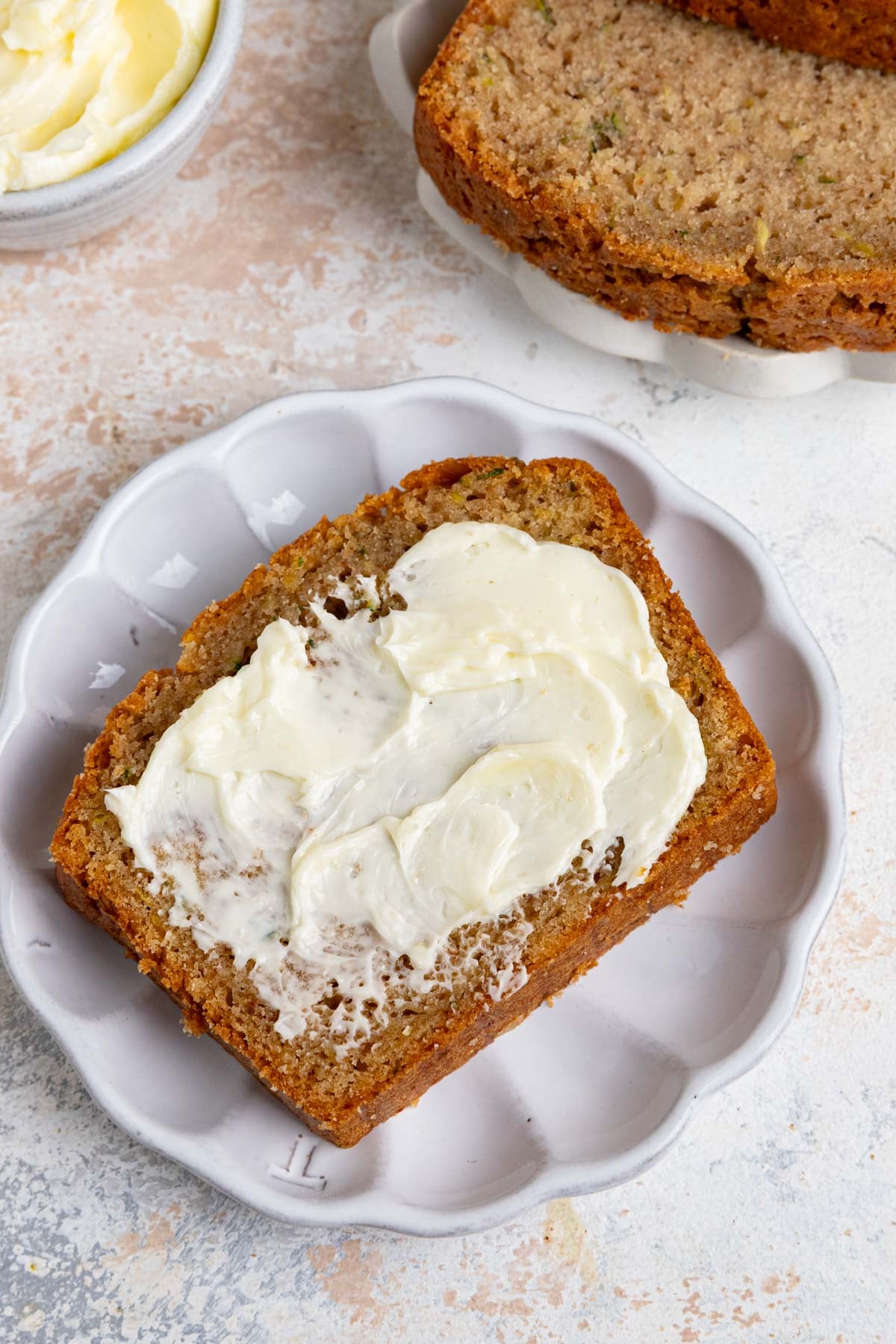 A slice of zucchini bread with butter spread on top sits on a white plate. Another slice of zucchini bread and a bowl of butter are visible in the background.