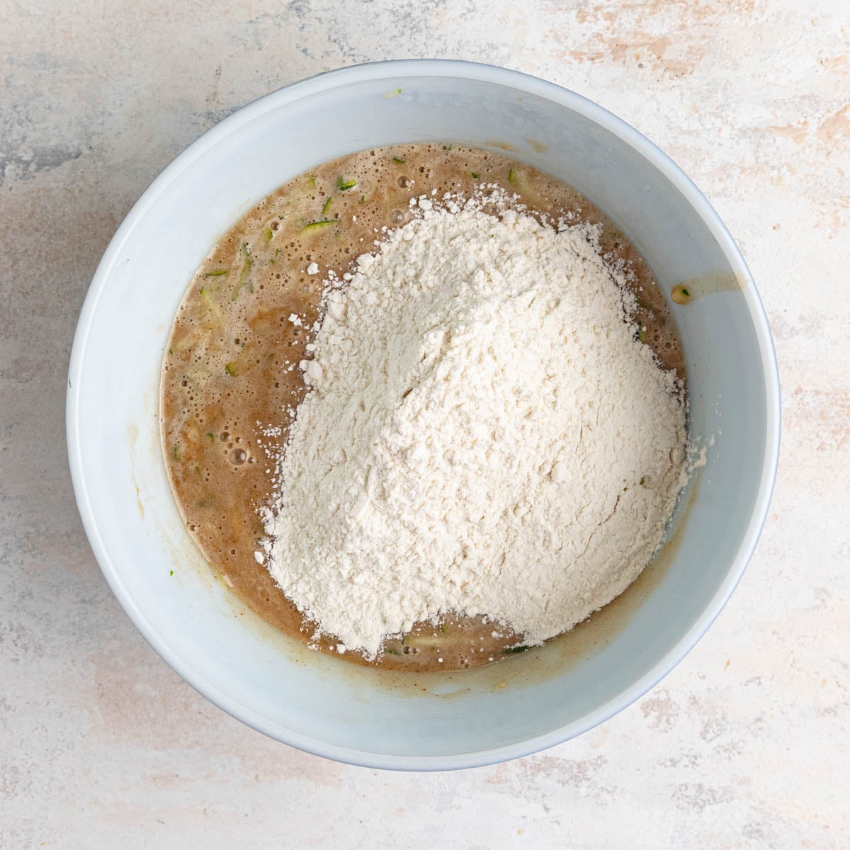 The zucchini bread batter in a white ceramic bowl with the flour flour mixture added and sitting on top