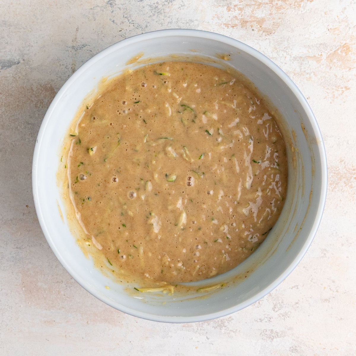 The fully mixed zucchini bread batter in a ceramic mixing bowl. The batter is light brown with flecks of green from the zucchini peel