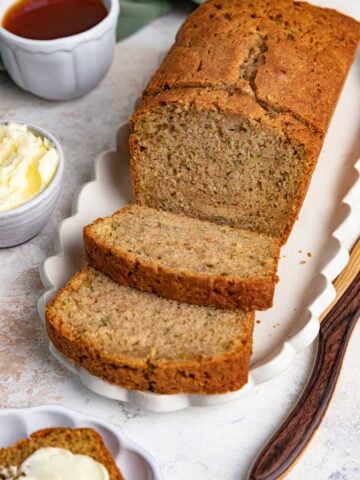A loaf of freshly baked zucchini bread is partially sliced on a white platter with a wooden knife. A small bowl of butter and a cup of tea are nearby, ready for serving. One slice of the moist zucchini bread is buttered on a separate plate.