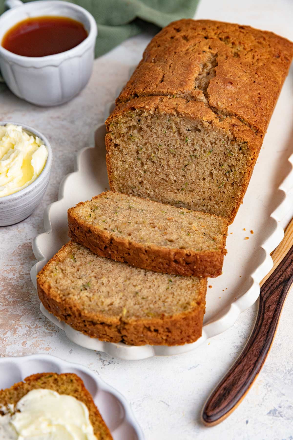 A loaf of freshly baked zucchini bread is partially sliced on a white platter with a wooden knife. A small bowl of butter and a cup of tea are nearby, ready for serving. One slice of the moist zucchini bread is buttered on a separate plate.