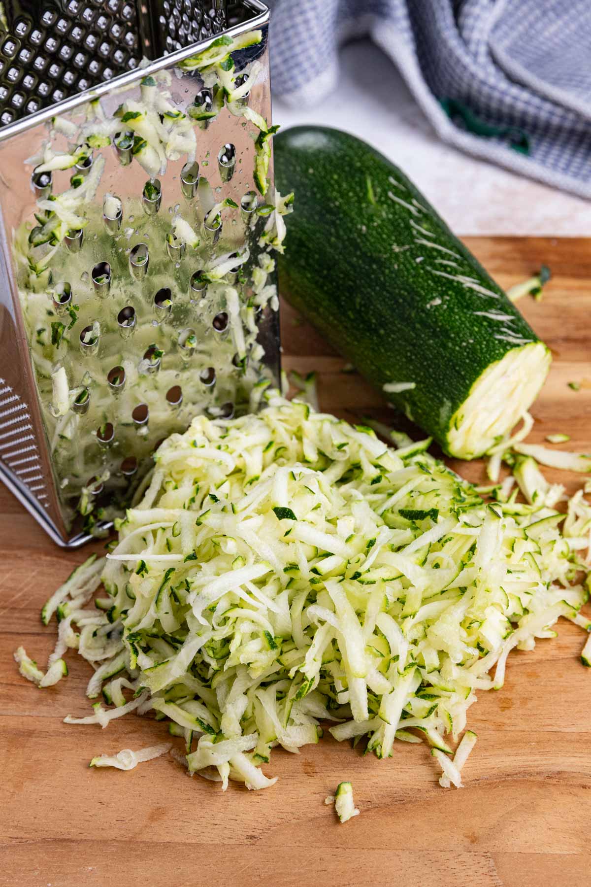 A metal box grater with shredded zucchini beside it sits on a wooden surface. A partially grated zucchini is next to the pile of shreds. A blue and white cloth is visible in the background.