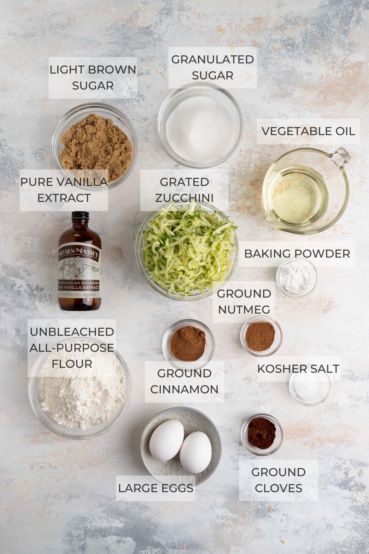 Top-down view of labeled baking ingredients in bowls and containers, including granulated sugar, vegetable oil, unbleached all-purpose flour, pure vanilla extract, grated zucchini, baking powder, ground cinnamon, ground nutmeg, ground cloves, kosher salt, and eggs.