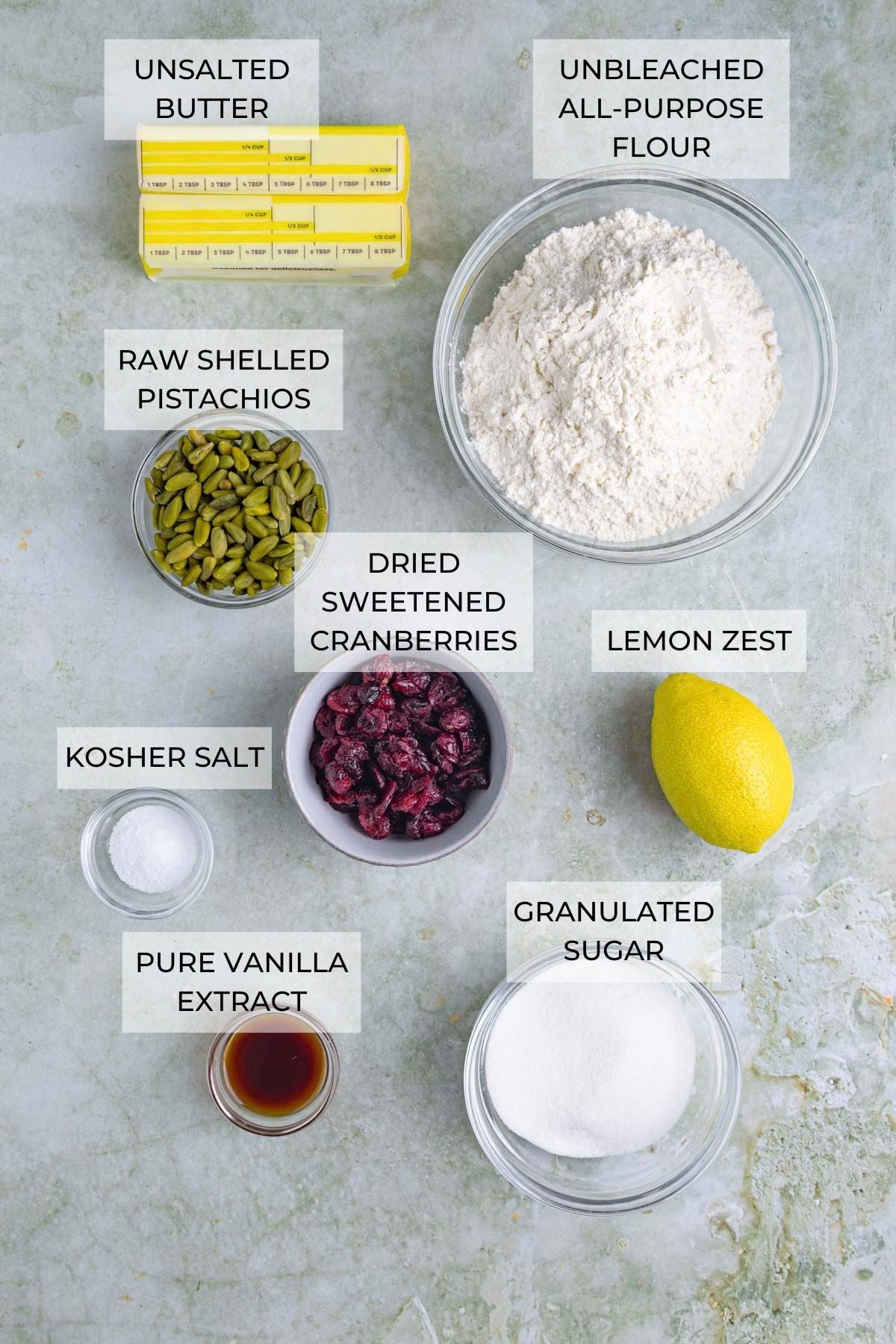 Cranberry pistachio shortbread cookie ingredients on a flat board - all-purpose flour, lemon zest (optional), granulated sugar, pure vanilla extract, kosher salt, dried sweetened cranberries, raw shelled pistachios, unsalted butter.