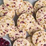 A pile of round cranberry and pistachio shortbread cookies studded with dried cranberries and pistachios, with small bowls of the dried cranberries and pistachios nearby