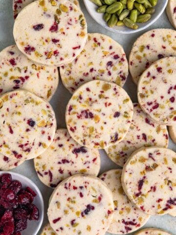 A pile of round cranberry and pistachio shortbread cookies studded with dried cranberries and pistachios, with small bowls of the dried cranberries and pistachios nearby
