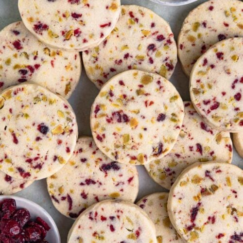 A pile of round cranberry and pistachio shortbread cookies studded with dried cranberries and pistachios, with small bowls of the dried cranberries and pistachios nearby