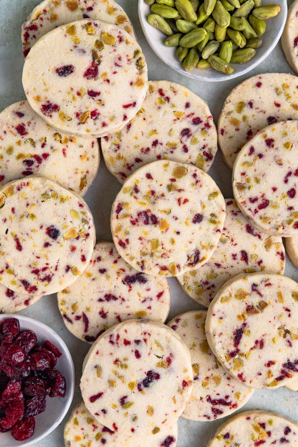 A pile of round cranberry and pistachio shortbread cookies studded with dried cranberries and pistachios, with small bowls of the dried cranberries and pistachios nearby