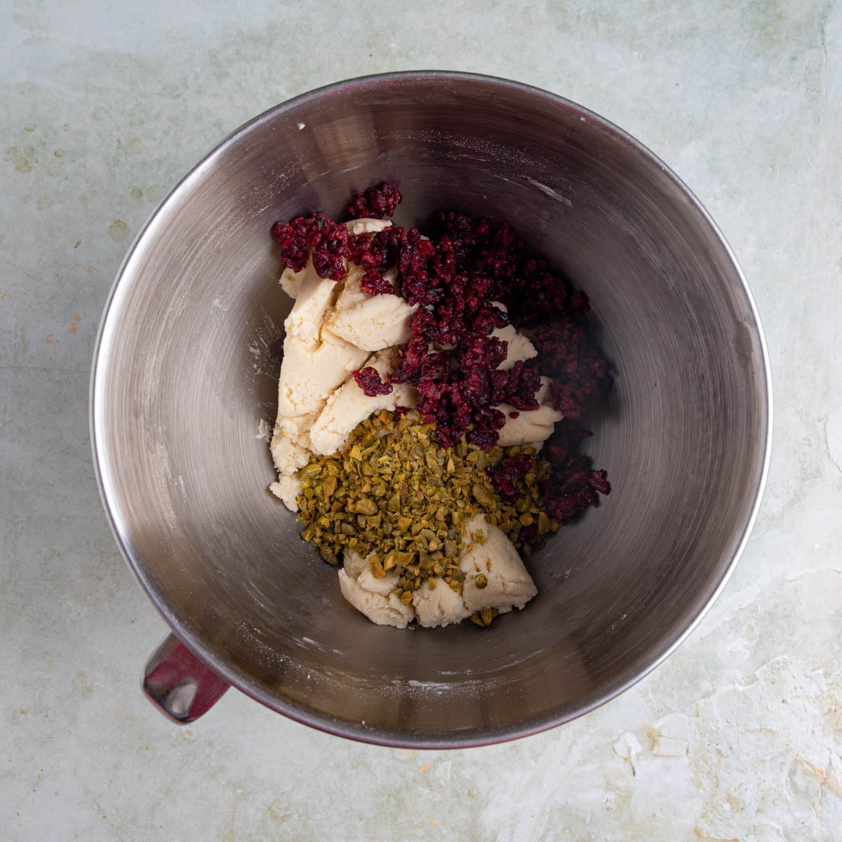 Shortbread cookie dough in stainless steel mixing bowl with chopped pistachios and chopped dried cranberries on top of the dough.