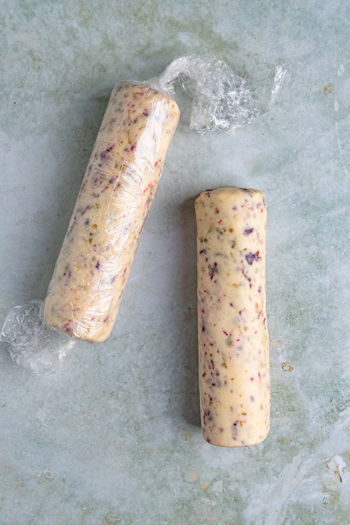 Two dough logs of cranberry pistachio shortbread cookie dough. One log is wrapped in plastic wrap.